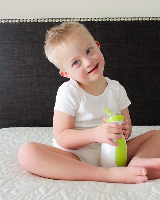Little boy with Down syndrome sitting on a bed, holding a Nosiboo Go portable electric nasal aspirator